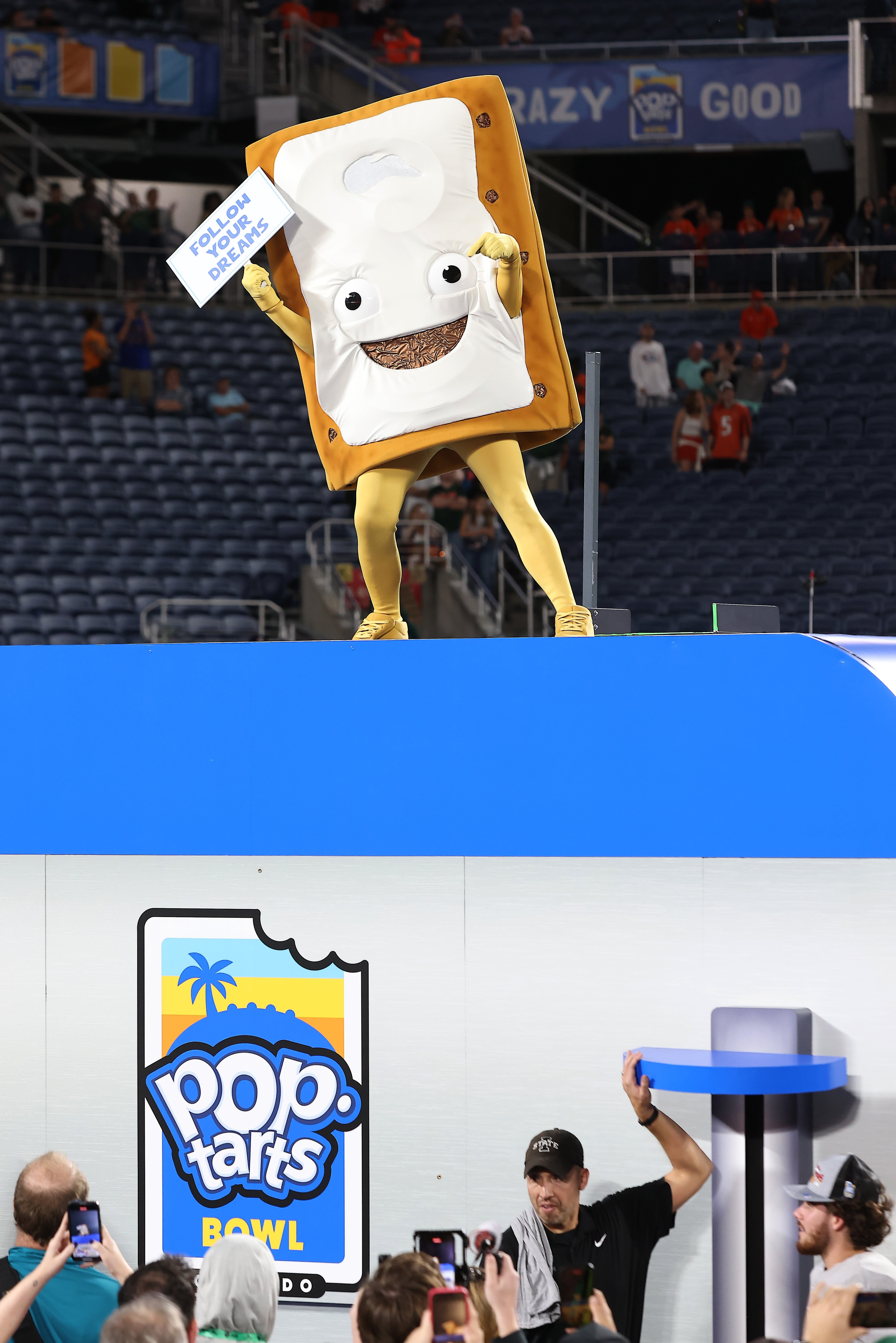 Mascot dressed arsenic a frosted pastry with arms holds a "Polish Off" motion astatine a sports event. Below, an jock prepares to statesman a challenge