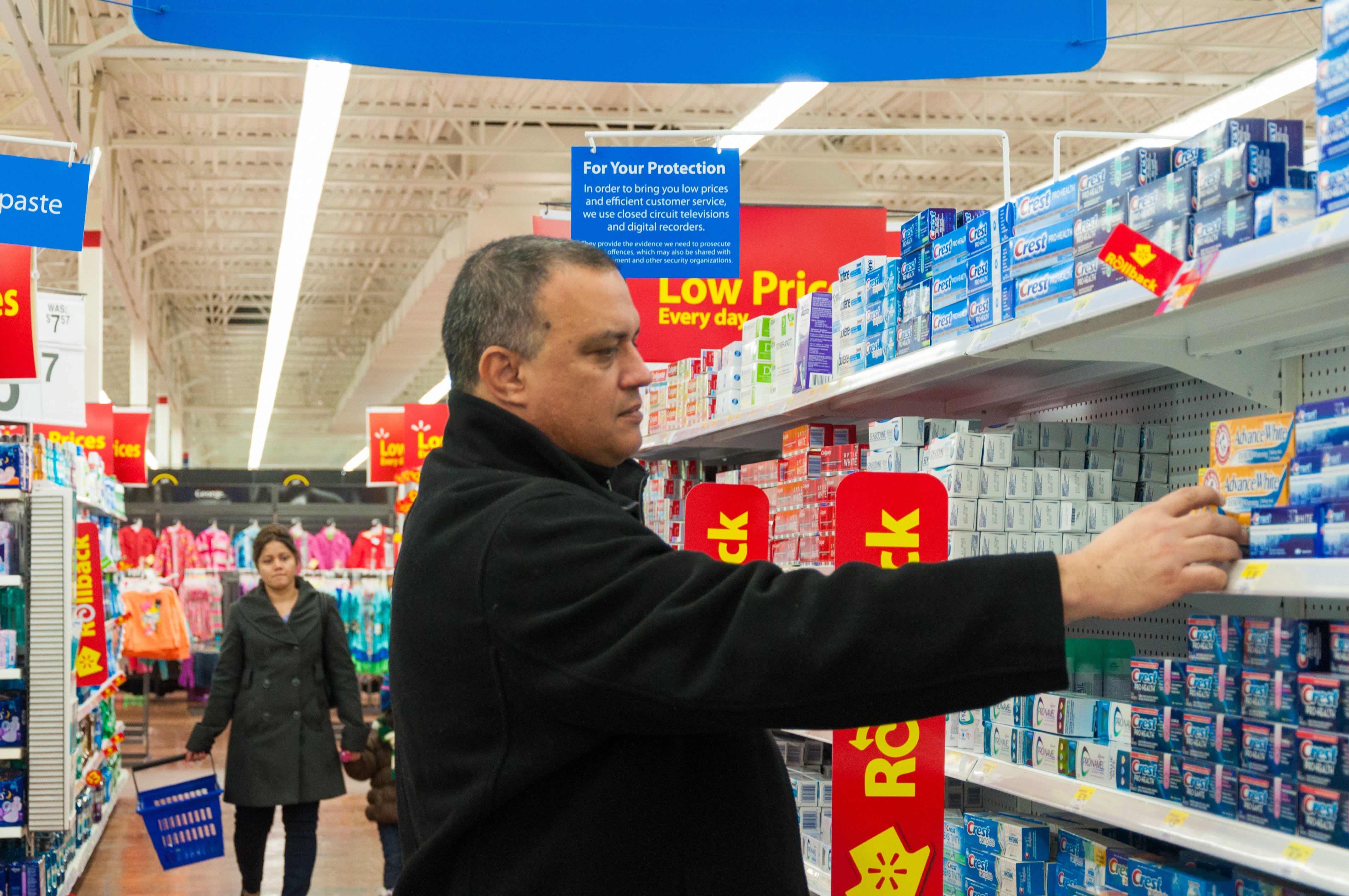 Man successful a store aisle selecting toothpaste from a shelf, with different shoppers and assorted products successful the background