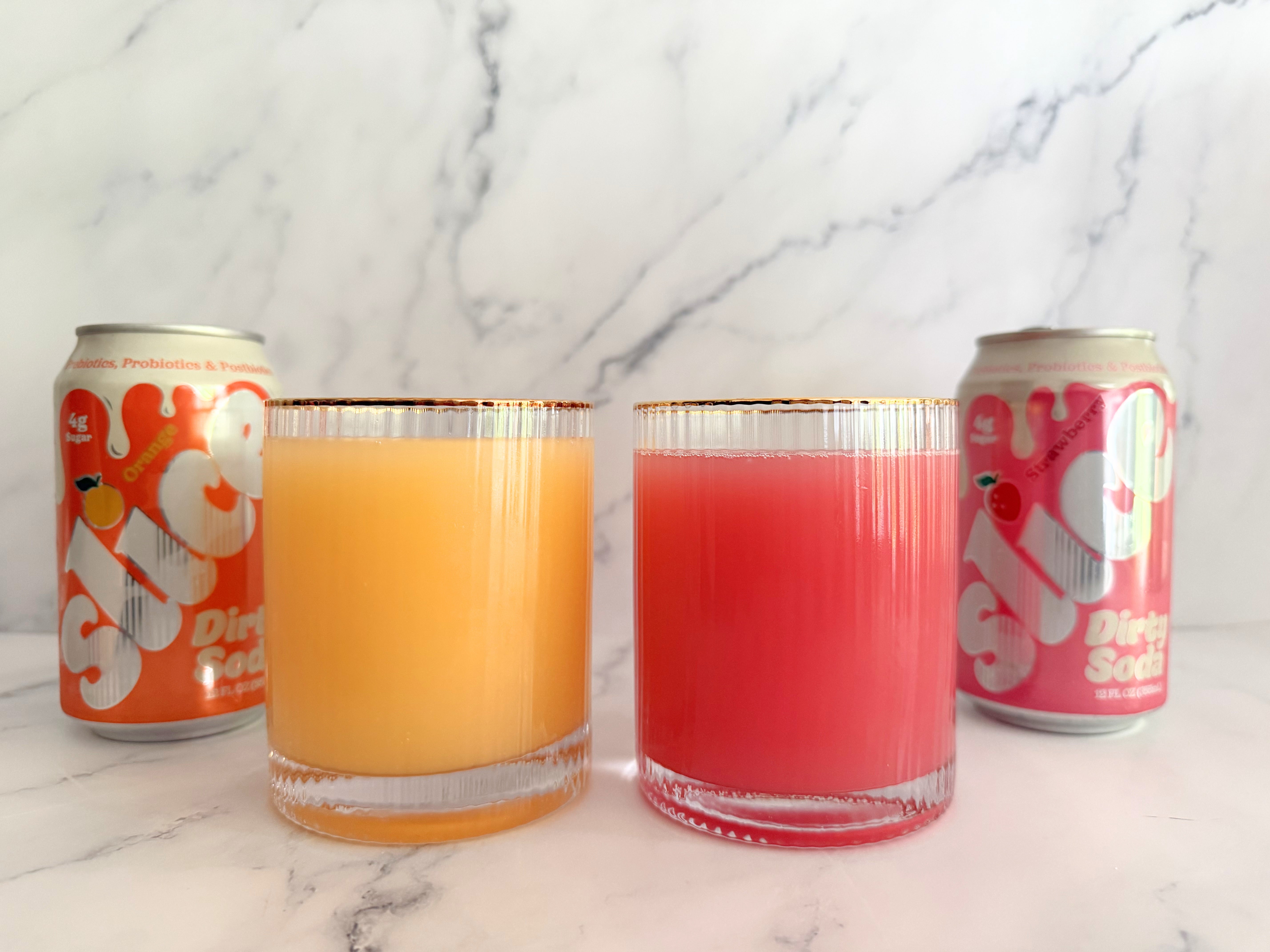 Two glasses of Slice Orange and Strawberry Dirty Sodas are in front of Slice soda cans with matching colors, set against a marble background