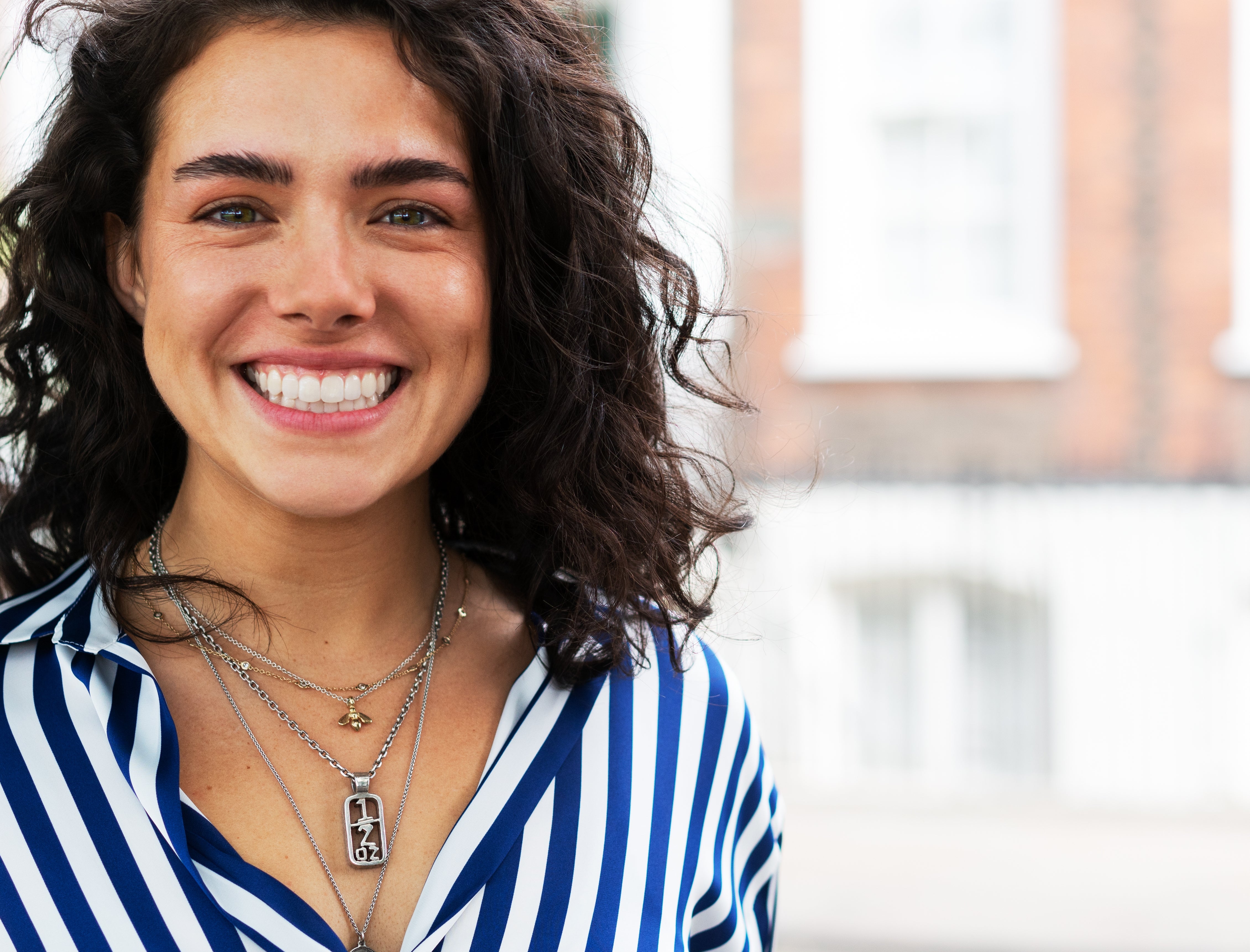 Person smiling outdoors, wearing a striped garment  and layered necklaces, with a blurred municipality  background