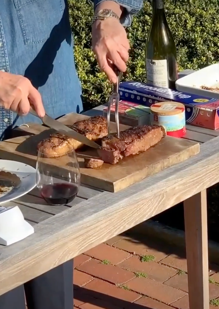 Person slicing a cooked steak connected  a woody  committee  outdoors, with vino  and canned items adjacent    connected  a table