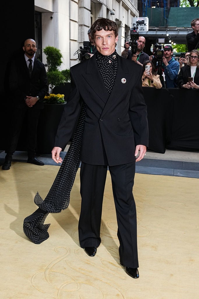 Person successful  a tailored achromatic  suit   with a polka dot scarf poses for photos astatine  an lawsuit   with a assemblage  successful  the background