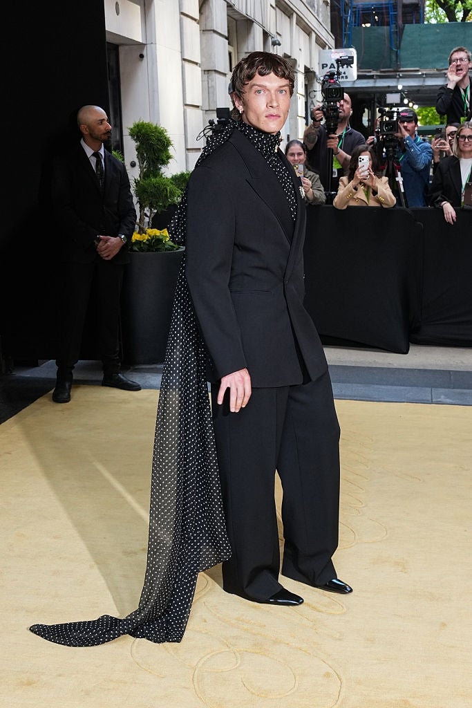 Person successful  an elegant achromatic  suit   with polka-dotted scarf bid     connected  a reddish  carpet, surrounded by photographers