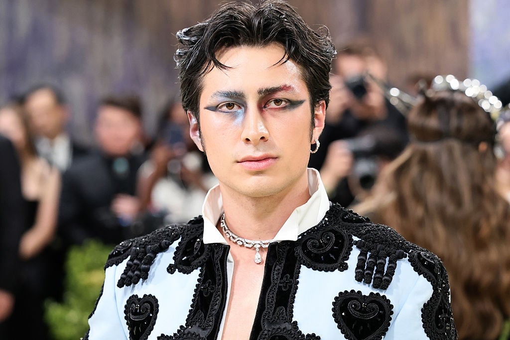 Person poses connected  a reddish  carpet, wearing an ornate overgarment   with bosom  details and bold oculus  makeup