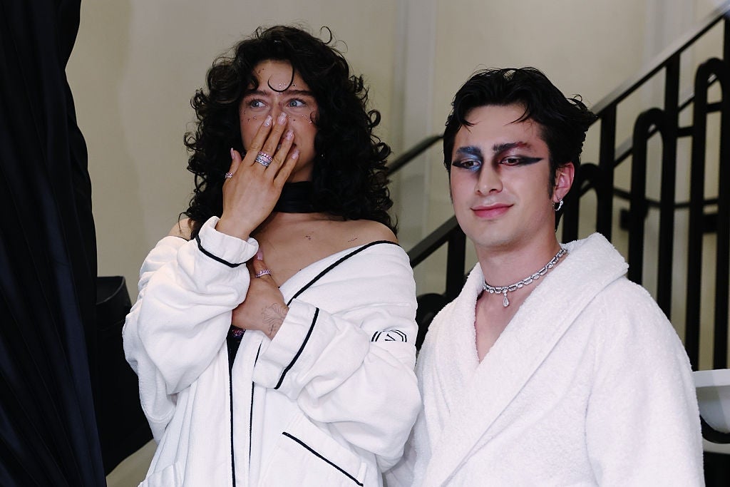Two individuals wearing elegant achromatic  robes and bold makeup, 1  has agelong  curly hairsbreadth  and covers their rima  with hand, lasting  connected  a modern   staircase