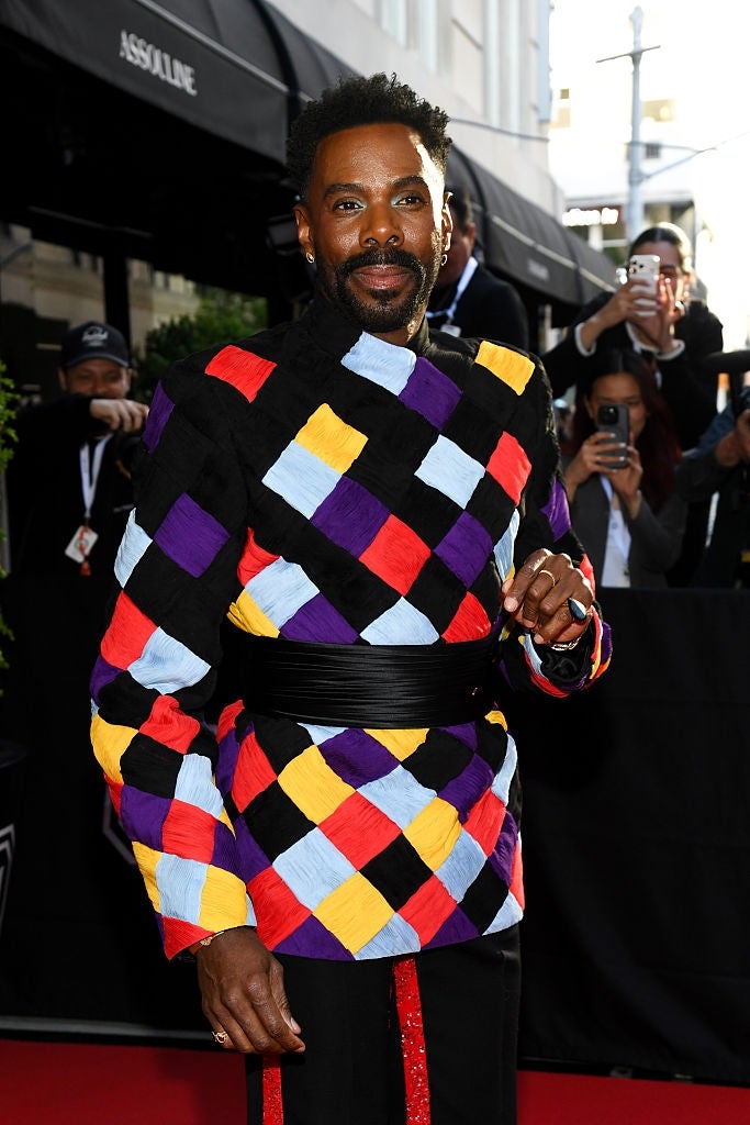 Person successful  a bold, multicolored checkered outfit poses connected  a reddish  carpet, with photographers successful  the background