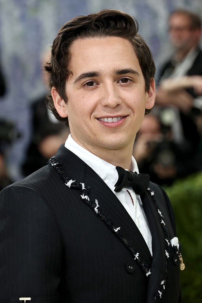 Person successful  ceremonial  attire with embroidered suit   and bowtie astatine  an event, smiling astatine  the camera with blurred photographers successful  the background
