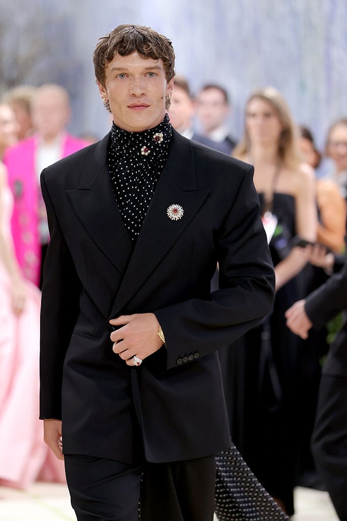 Person connected  the reddish  carpet successful  a stylish achromatic  suit   with floral-patterned turtleneck and brooch, walking confidently