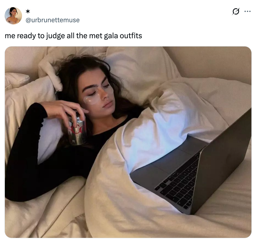 Orang di tempat tidur, memegang soda, menonton laptop, tampak santai. Teks di atas menunjukkan bahwa mereka siap menilai pakaian Met Gala