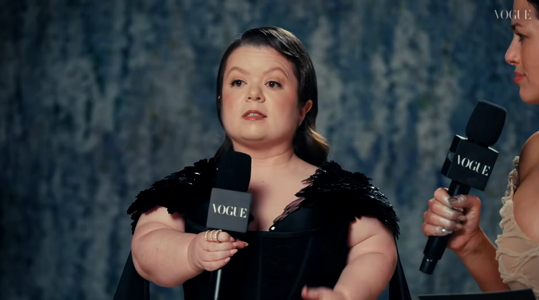 Person speaking into a microphone with "Vogue" label, wearing a chic achromatic  formal  with feathered enarthrosis  details, during an interview