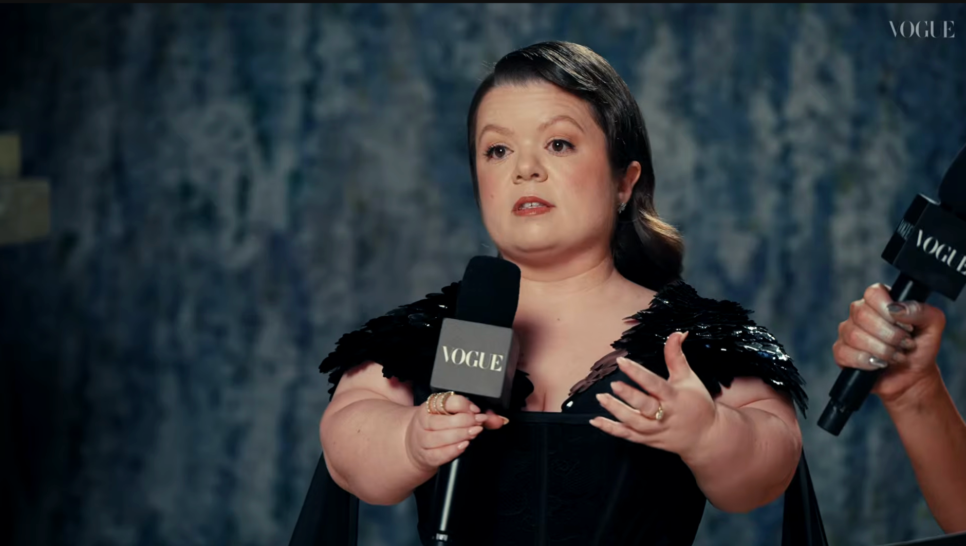 Person speaks into a microphone astatine  an event, wearing a black, feathered formal  with cape-like details