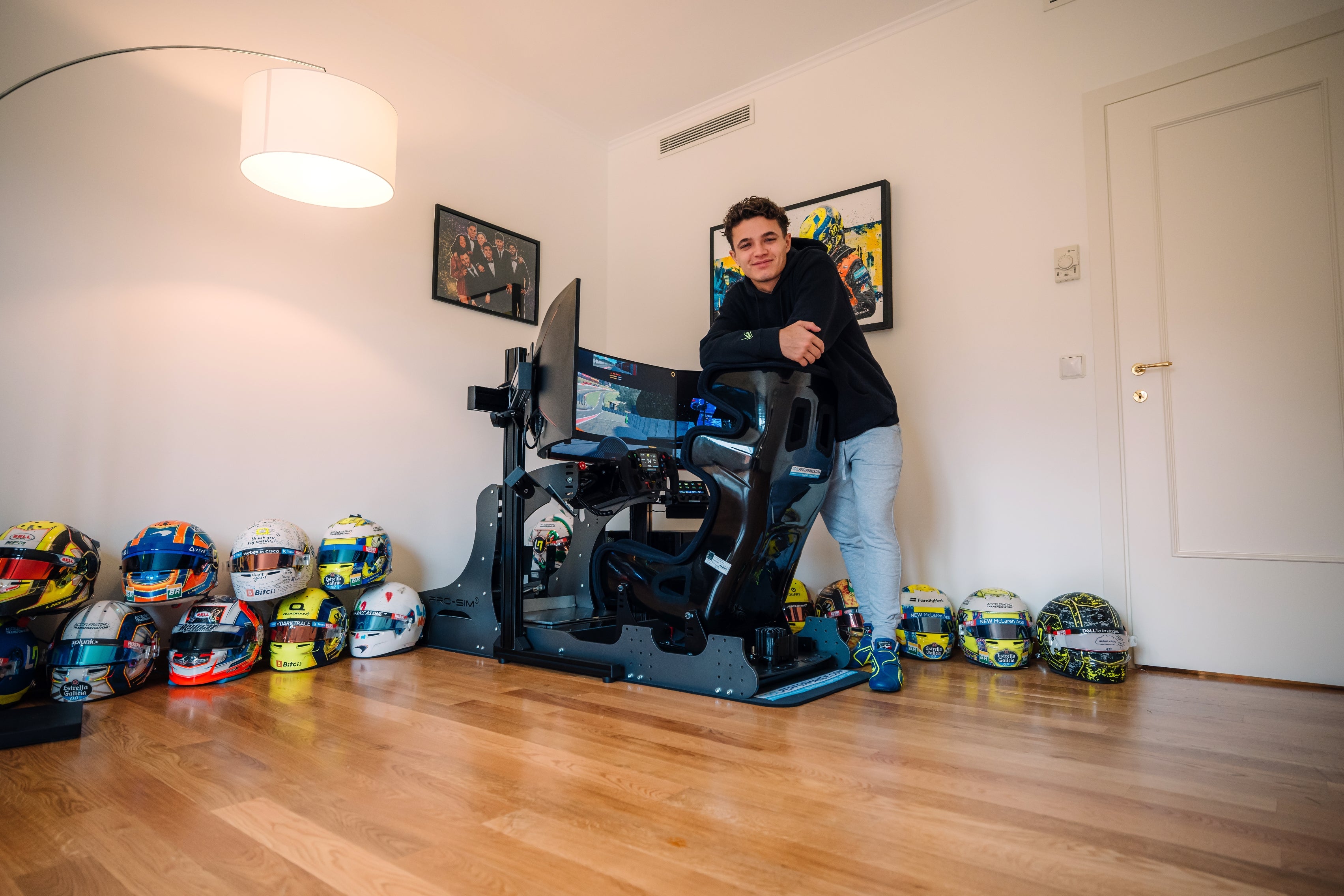 Person leaning on a racing simulator in a room with multiple racing helmets on the floor and framed racing memorabilia on the walls