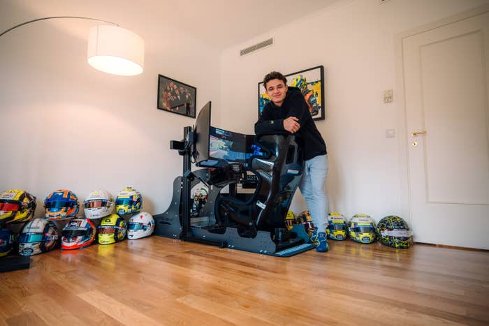 Person leaning on a racing simulator in a room with multiple racing helmets on the floor and framed racing memorabilia on the walls