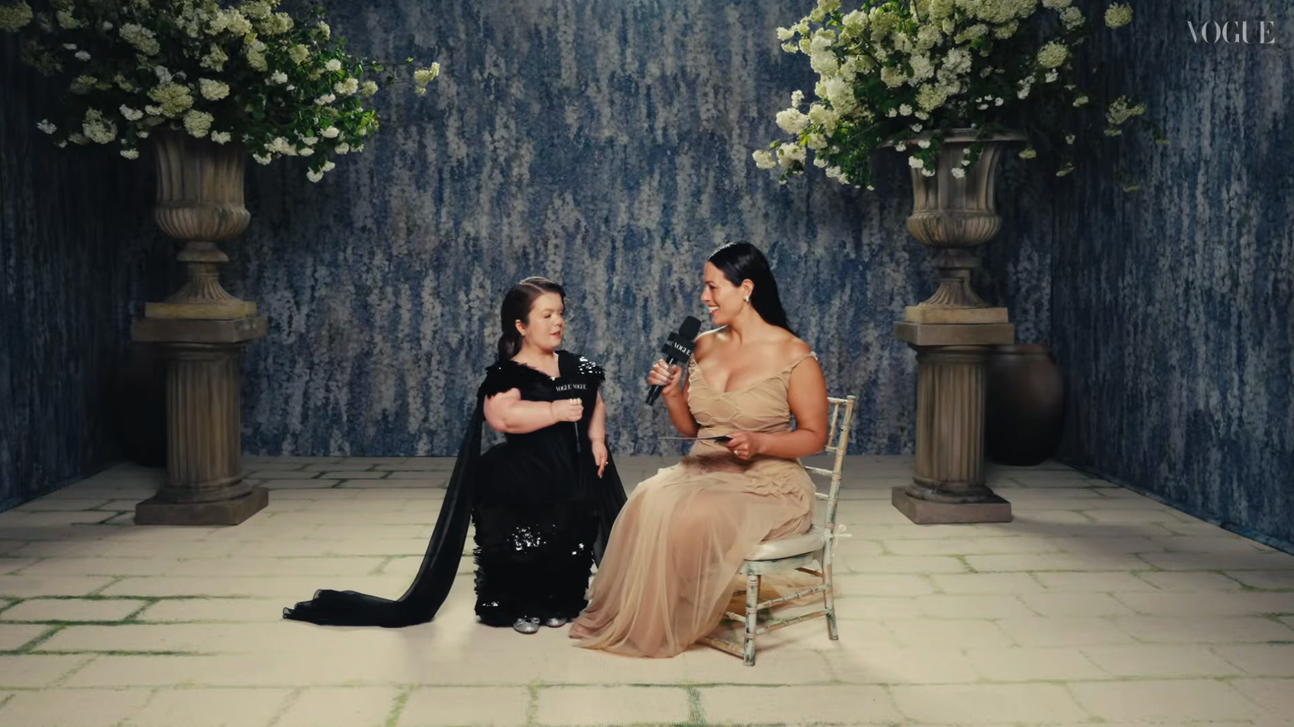 Two women sitting, 1  successful  a achromatic  gown and the different   successful  an elegant beige dress, engaged successful  speech  amid a floral backdrop