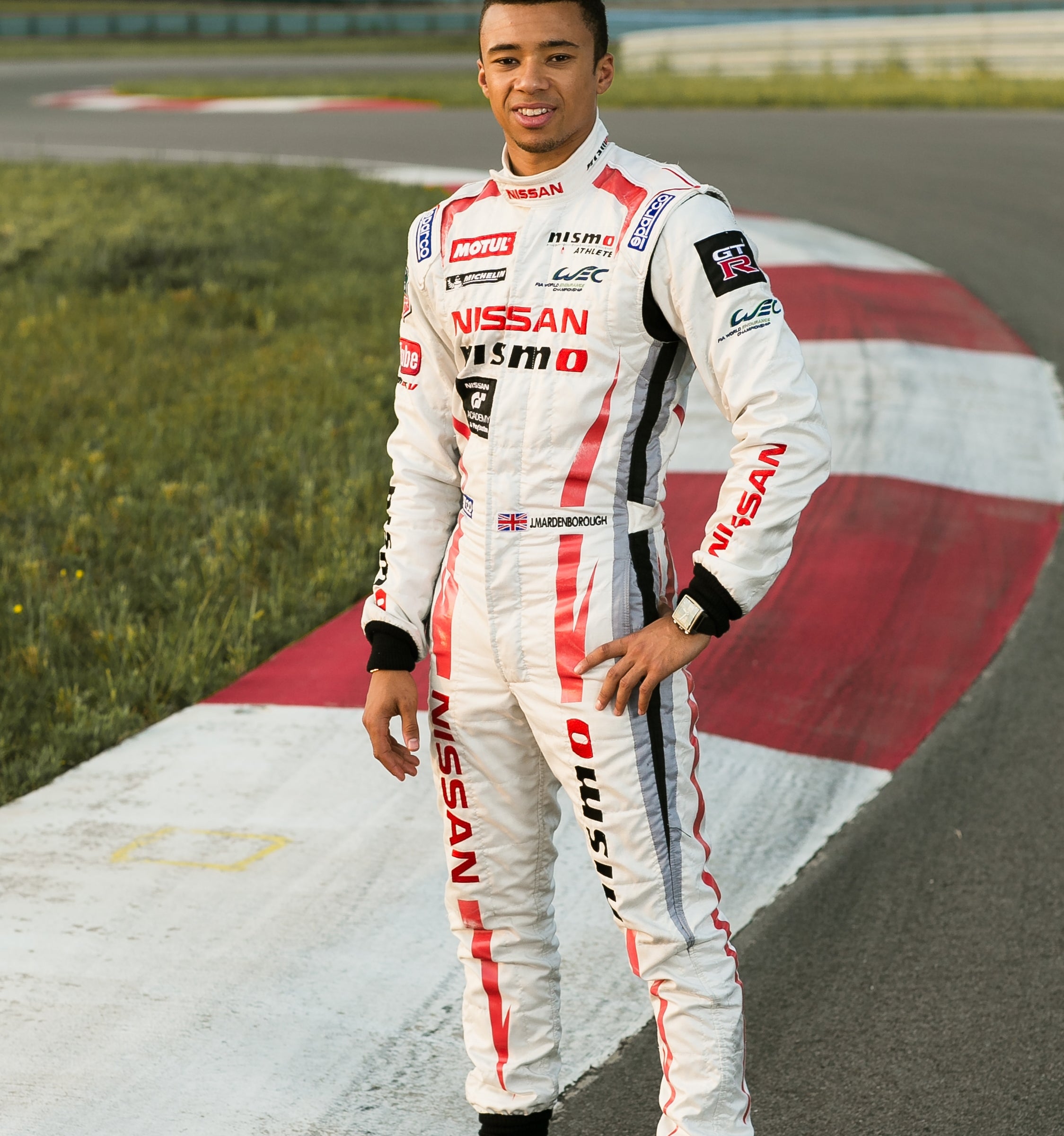 Race car driver in a branded jumpsuit standing on a racetrack, smiling at the camera