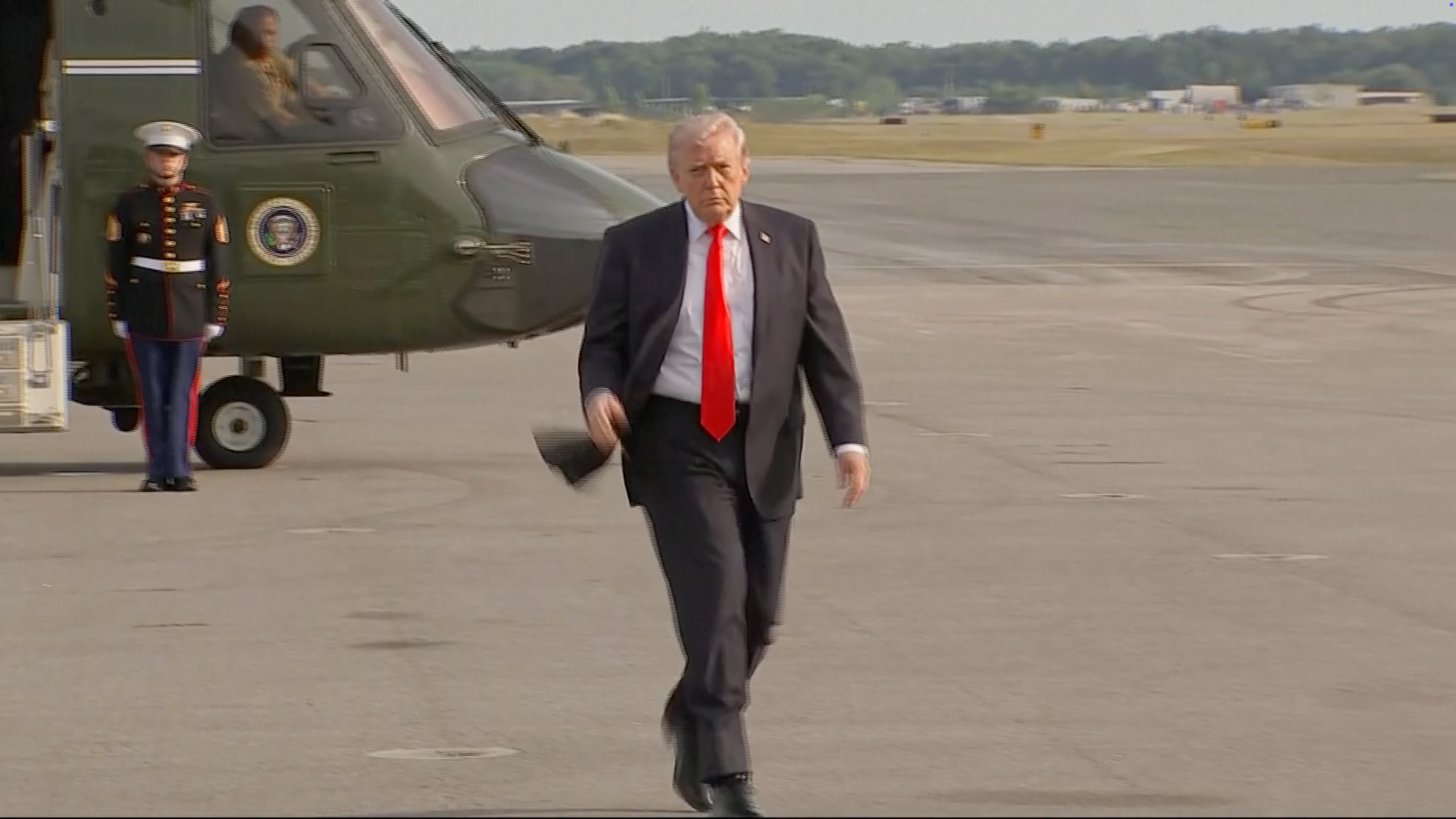 A antheral   successful  a suit   with a reddish  necktie  walks connected  an airfield adjacent   a helicopter, accompanied by a Marine guard