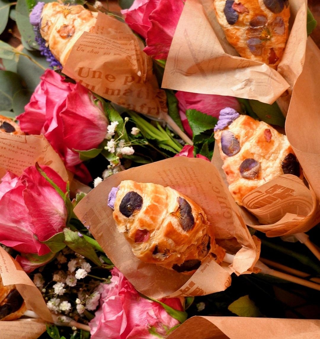 Bouquet of croissants wrapped in paper alongside pink roses and greenery, creating a pastry and floral arrangement