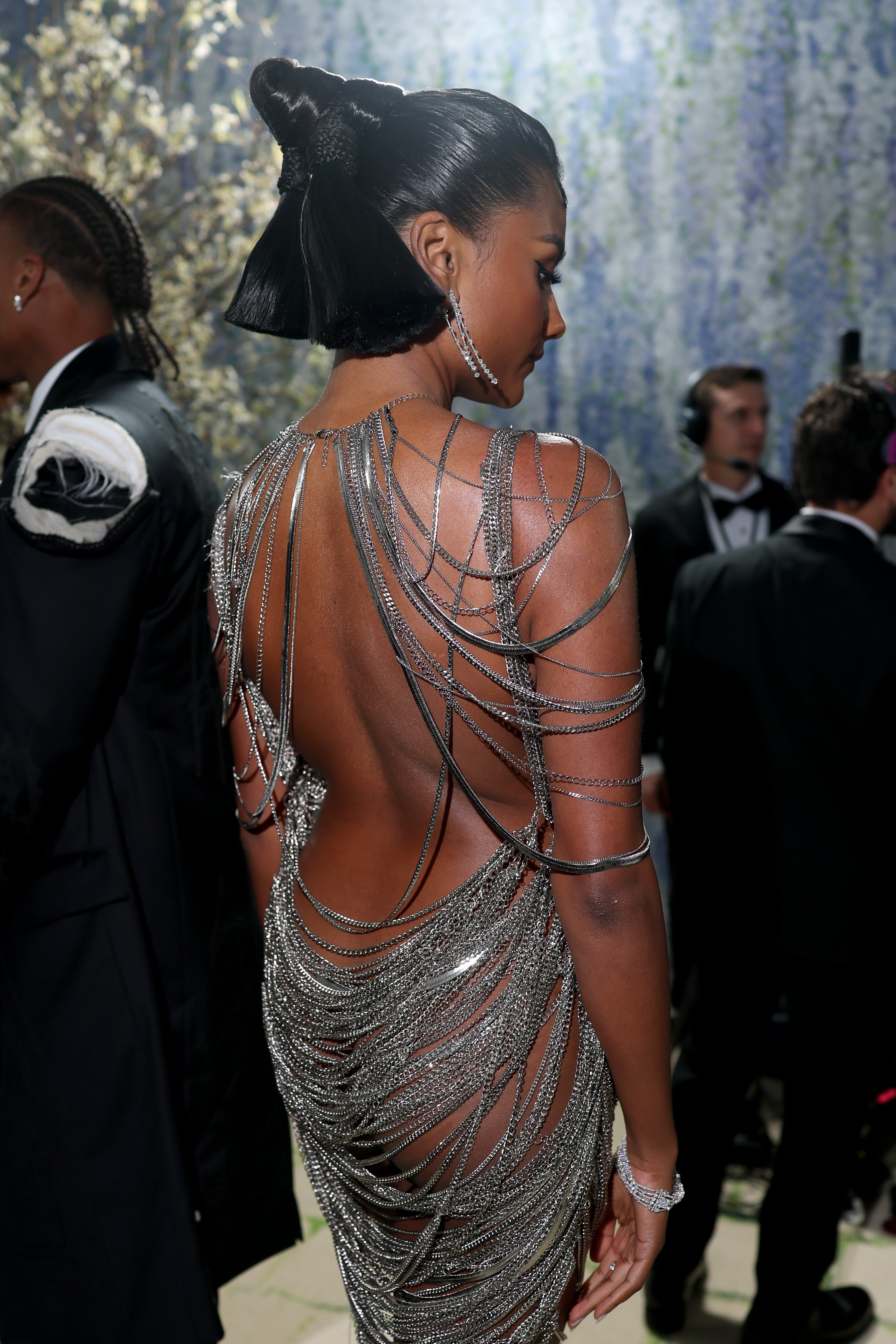 Person successful  elegant backless gown draped with shimmering chains, astatine  an event. Hair styled successful  a sleek updo. Others successful  ceremonial  deterioration  successful  the background