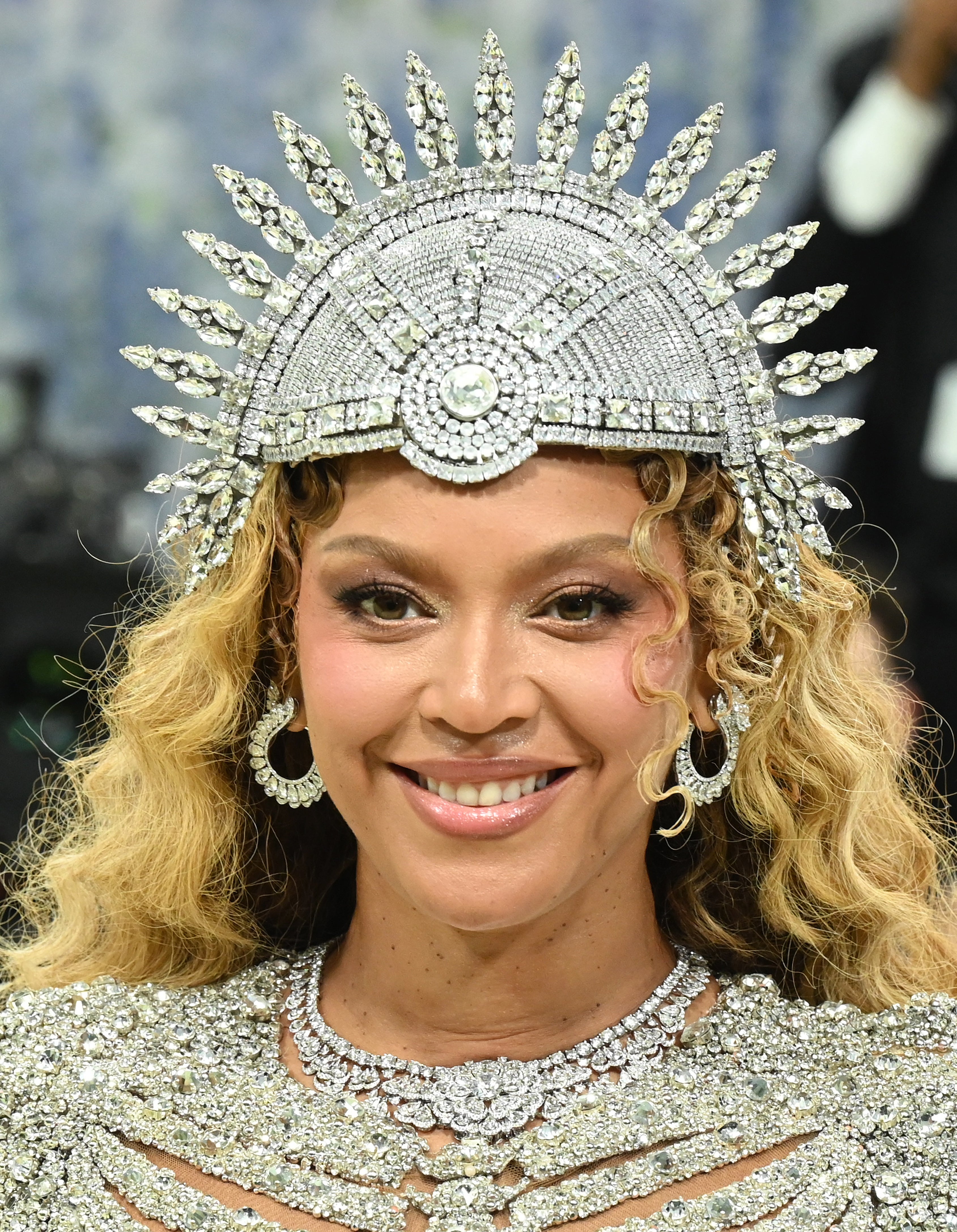 Person wearing an ornate, jewel-encrusted headdress with a spiked design, smiling astatine  the camera