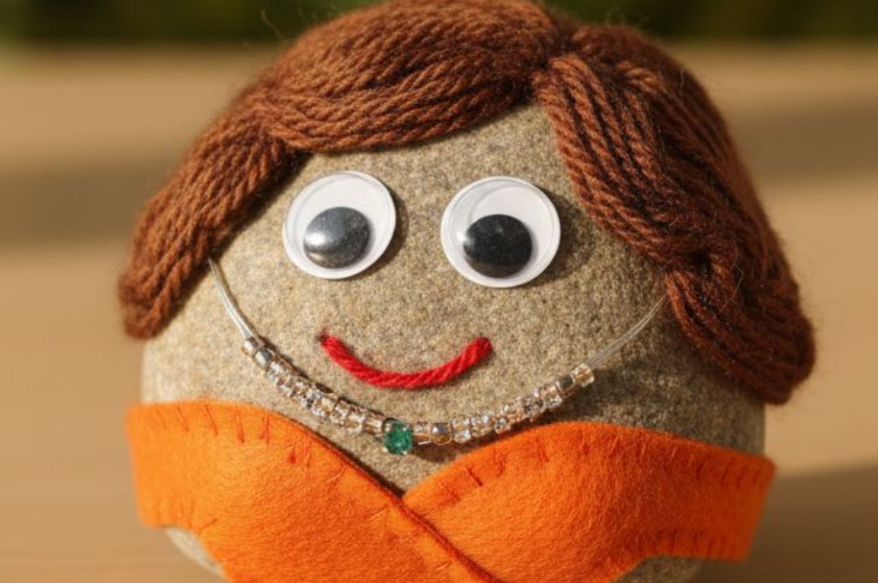 Toy rock with googly eyes, yarn hair, and orange felt dress, smiling on a wooden surface