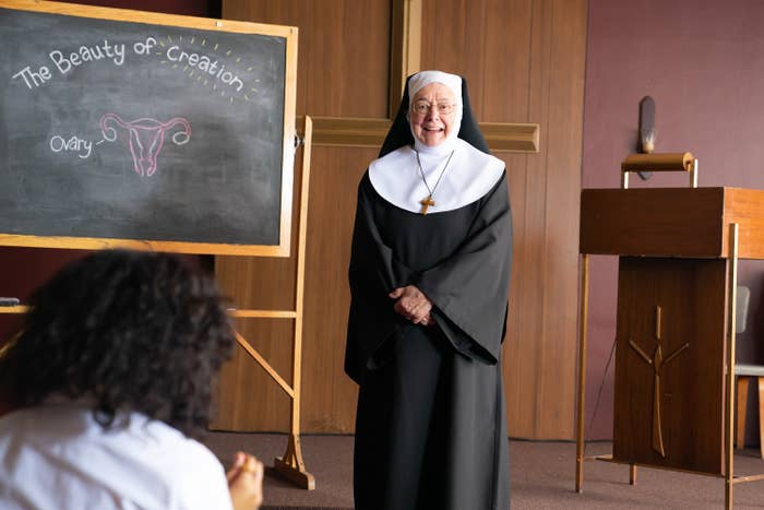A smiling nun stands by a chalkboard with a uterus drawing and text "The Beauty of Creation" in a classroom setting