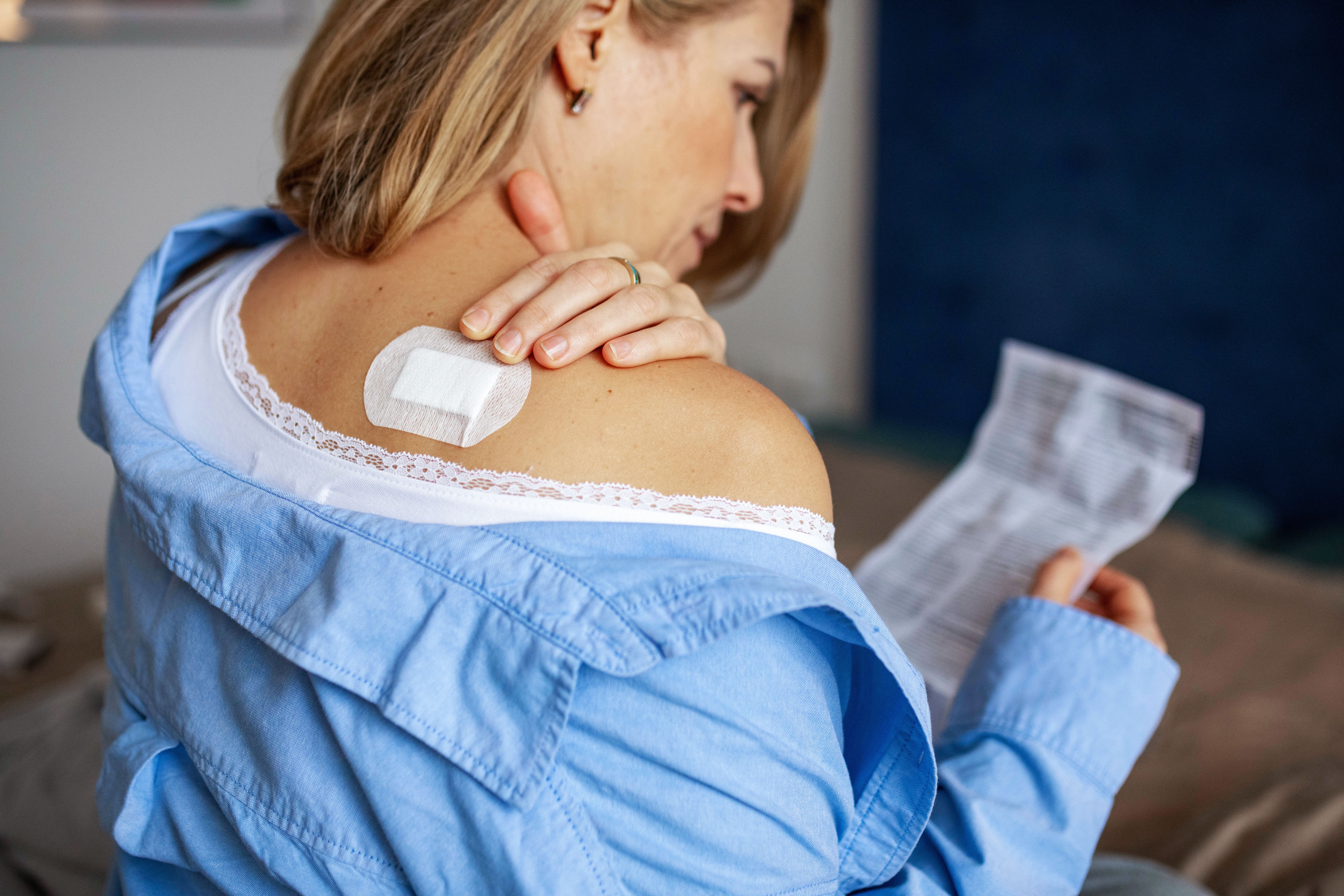 Woman with a contraceptive patch on shoulder reads instructions in bedroom setting, showcasing a method of birth control
