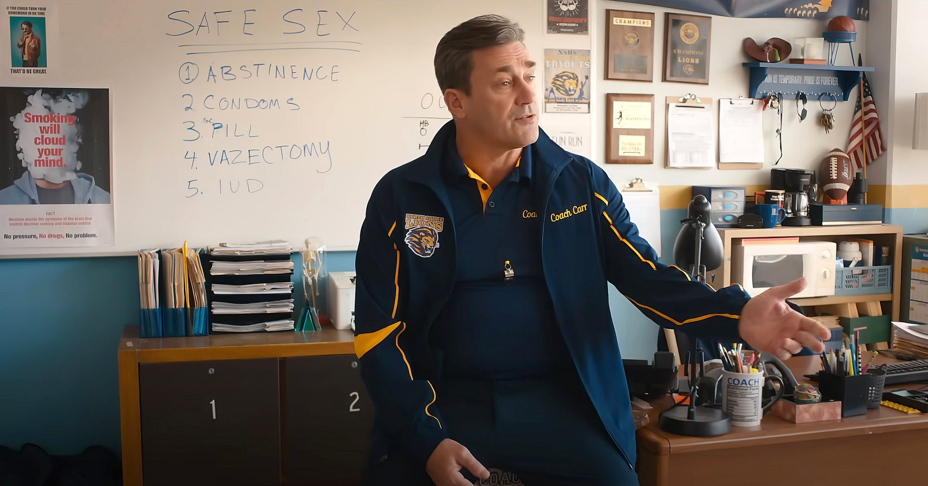 A coach sits in an office beside a board listing safe sex methods, discussing the topic