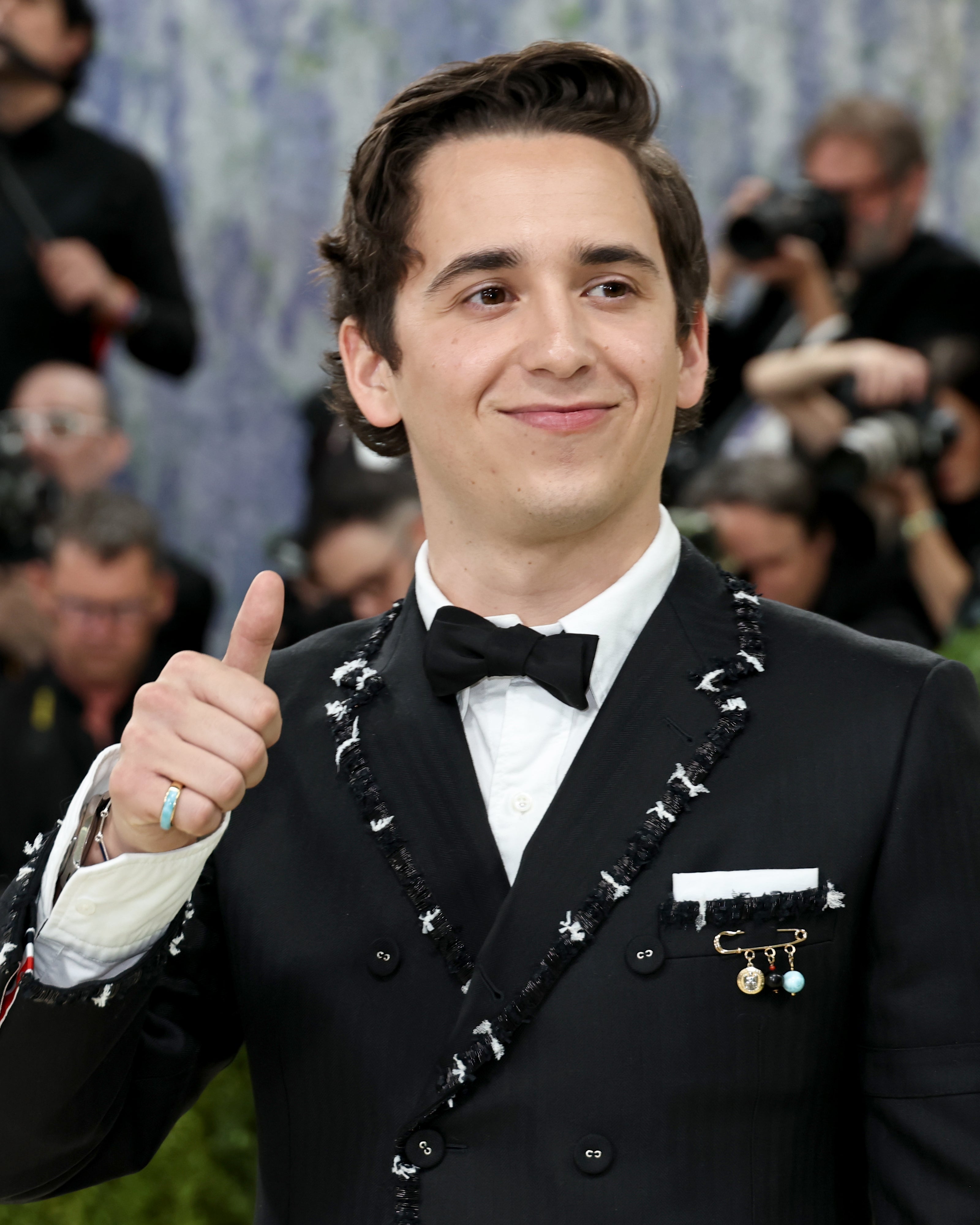 Person successful  a achromatic  tuxedo with decorative edges, bow tie, and brooches gives a thumbs-up connected  a ceremonial  lawsuit   carpet with photographers successful  the background