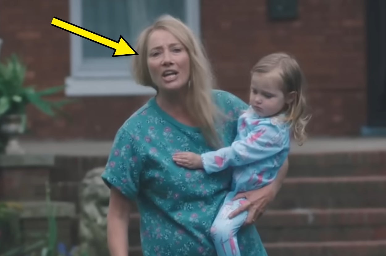 A woman holding a child stands outside a building, engaging in conversation. She is dressed casually