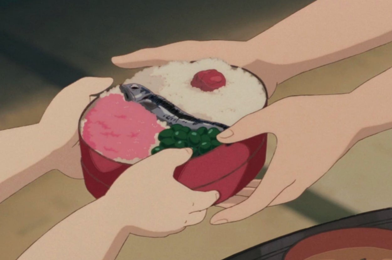 Two hands present a bowl of rice with a fish, garnish, and topping to another pair of hands