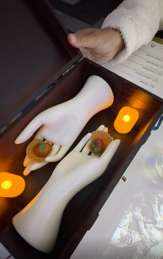 Decorative hands in a box with candles and pastries, next to a person's arm holding the lid