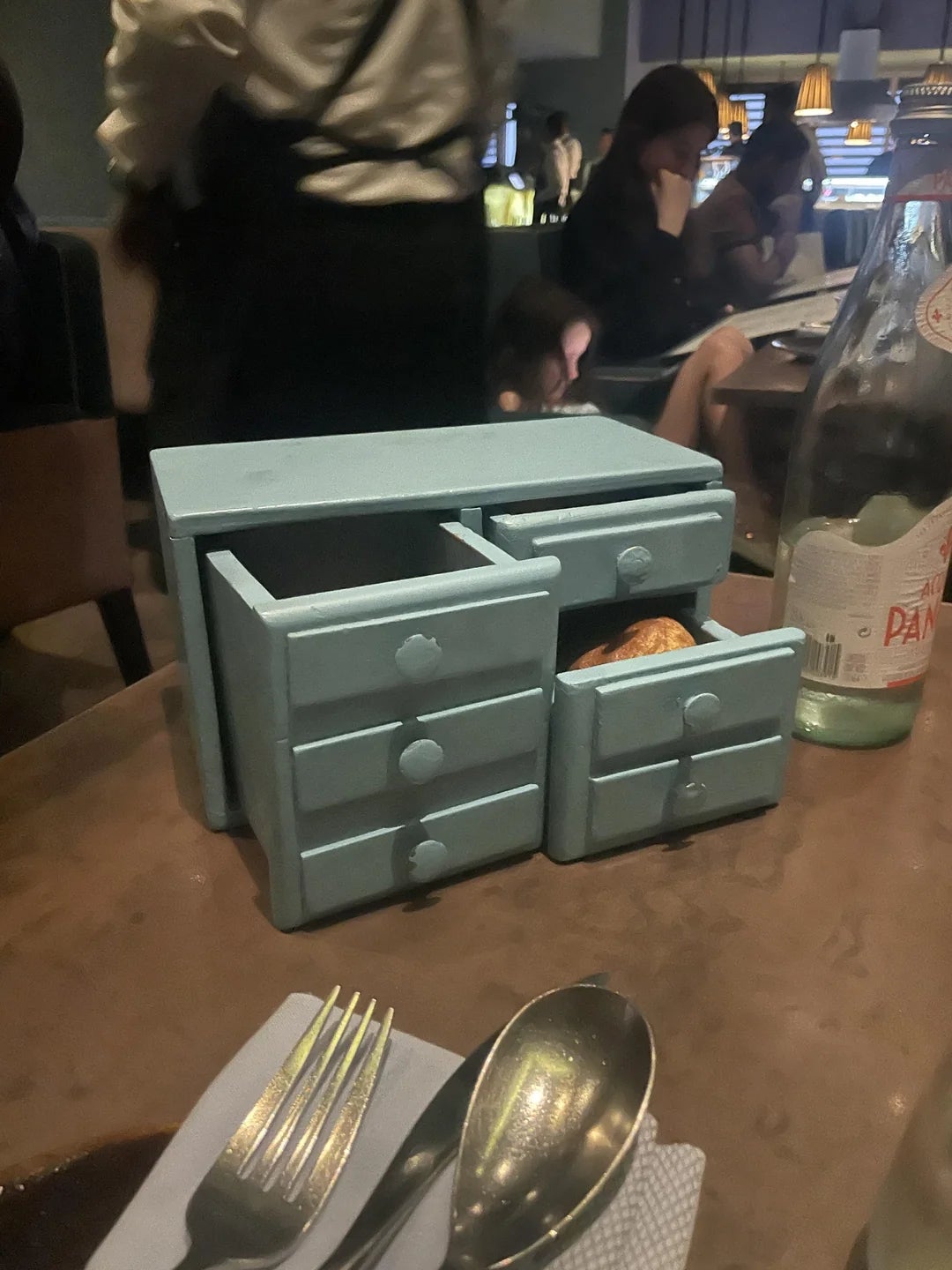 Miniature blue dresser with drawers holds a croissant, placed on a restaurant table along with cutlery and a mineral water bottle