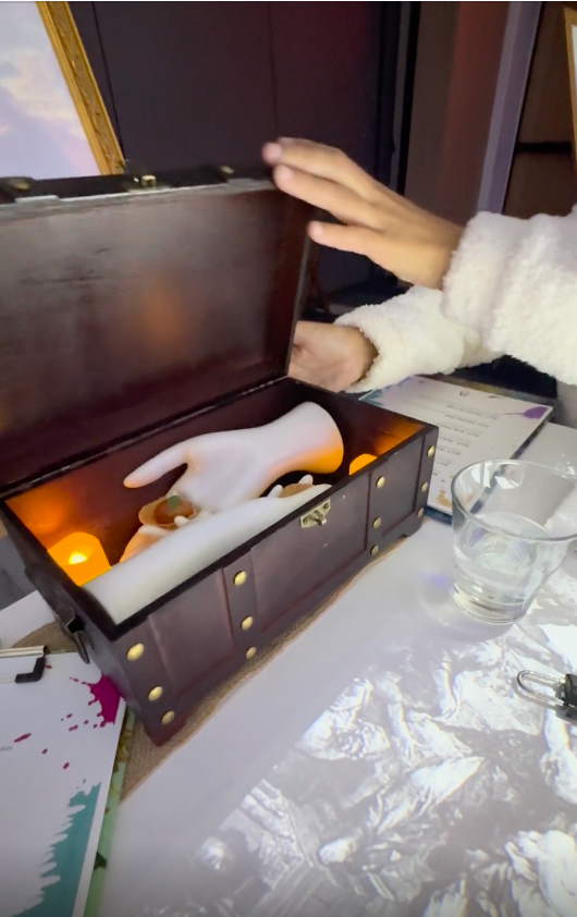 A person in a fluffy sweater opens a chest revealing a white statue hand, candles, and art supplies on the table