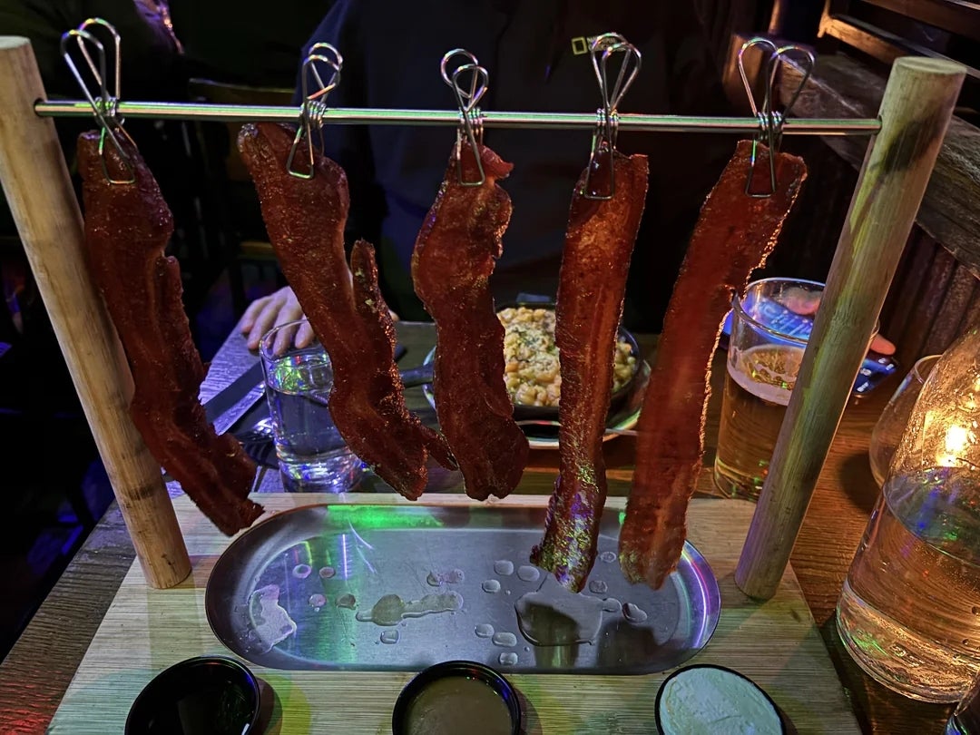 Bacon strips hanging on hooks on a rack over a tray, with small bowls of dipping sauces on the side