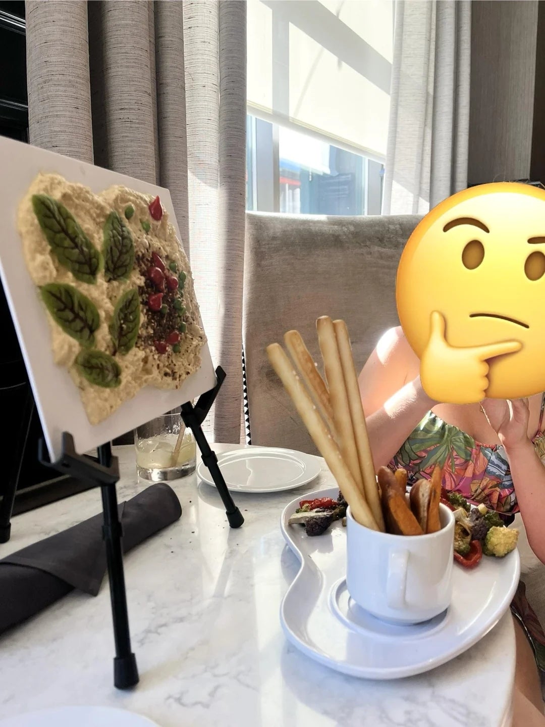 Person with a thinking emoji over their face at a restaurant. Table displays a creative dish resembling art, with breadsticks and sides