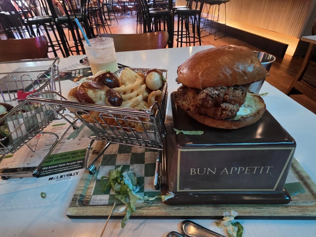 Fried chicken sandwich on a square pedestal plate with a sign reading "Bun Appetit," served with waffle fries and a drink in a burger-themed restaurant