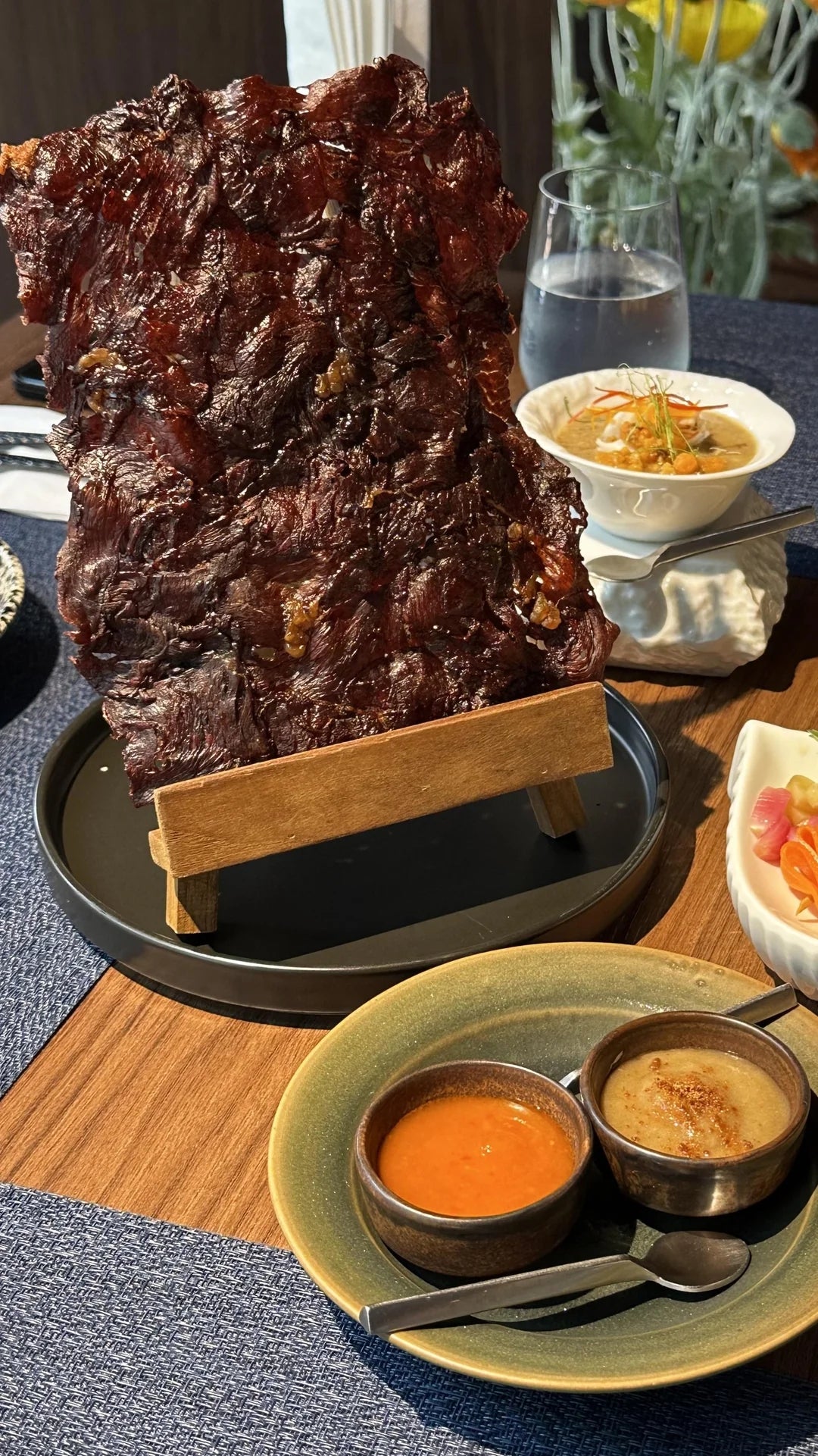 Large piece of grilled beef jerky displayed on a stand, accompanied by sauces, a bowl of soup, and a salad on a dining table