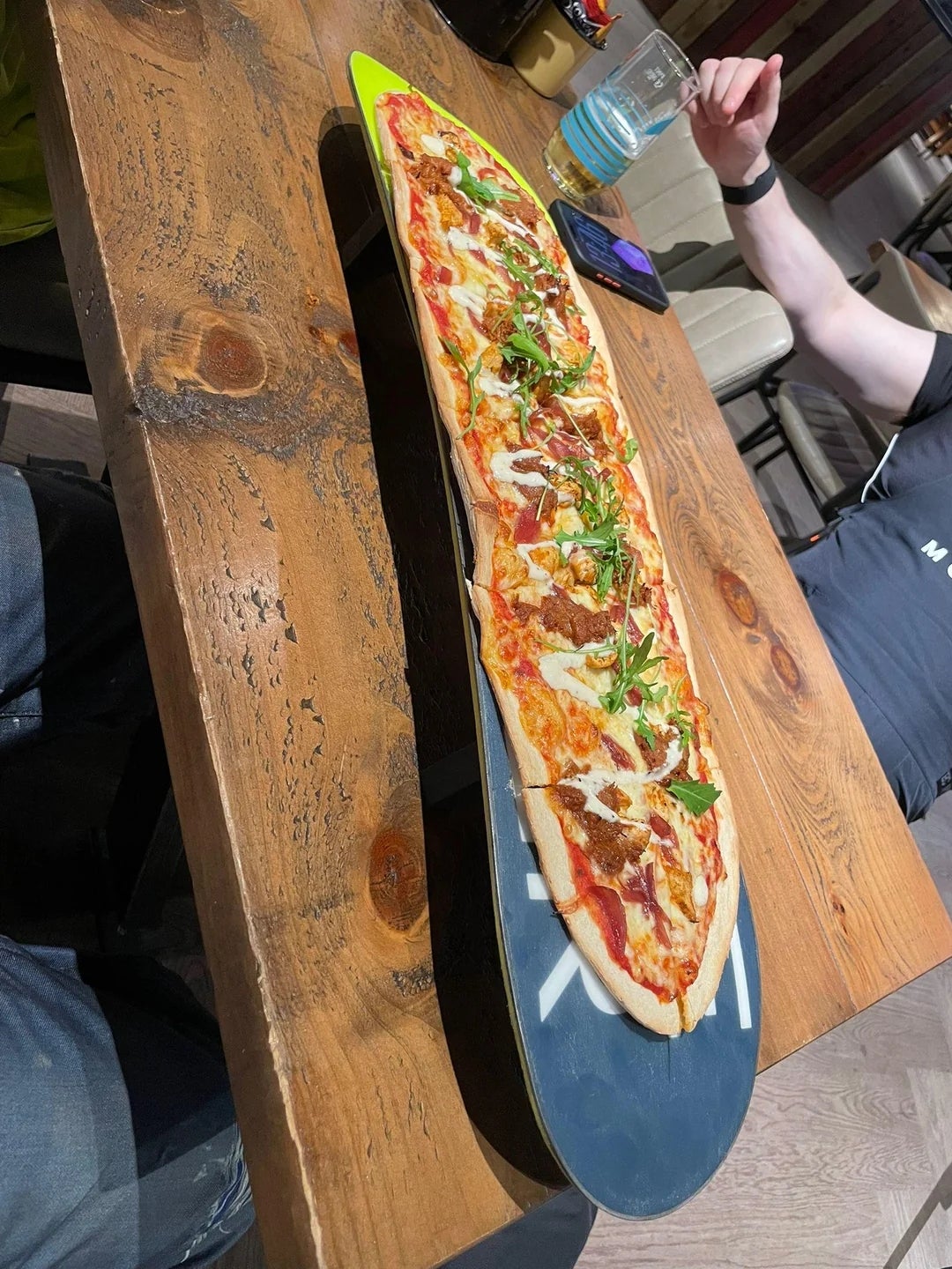 Pizza served on a skateboard as a platter, topped with arugula and other ingredients, on a wooden table