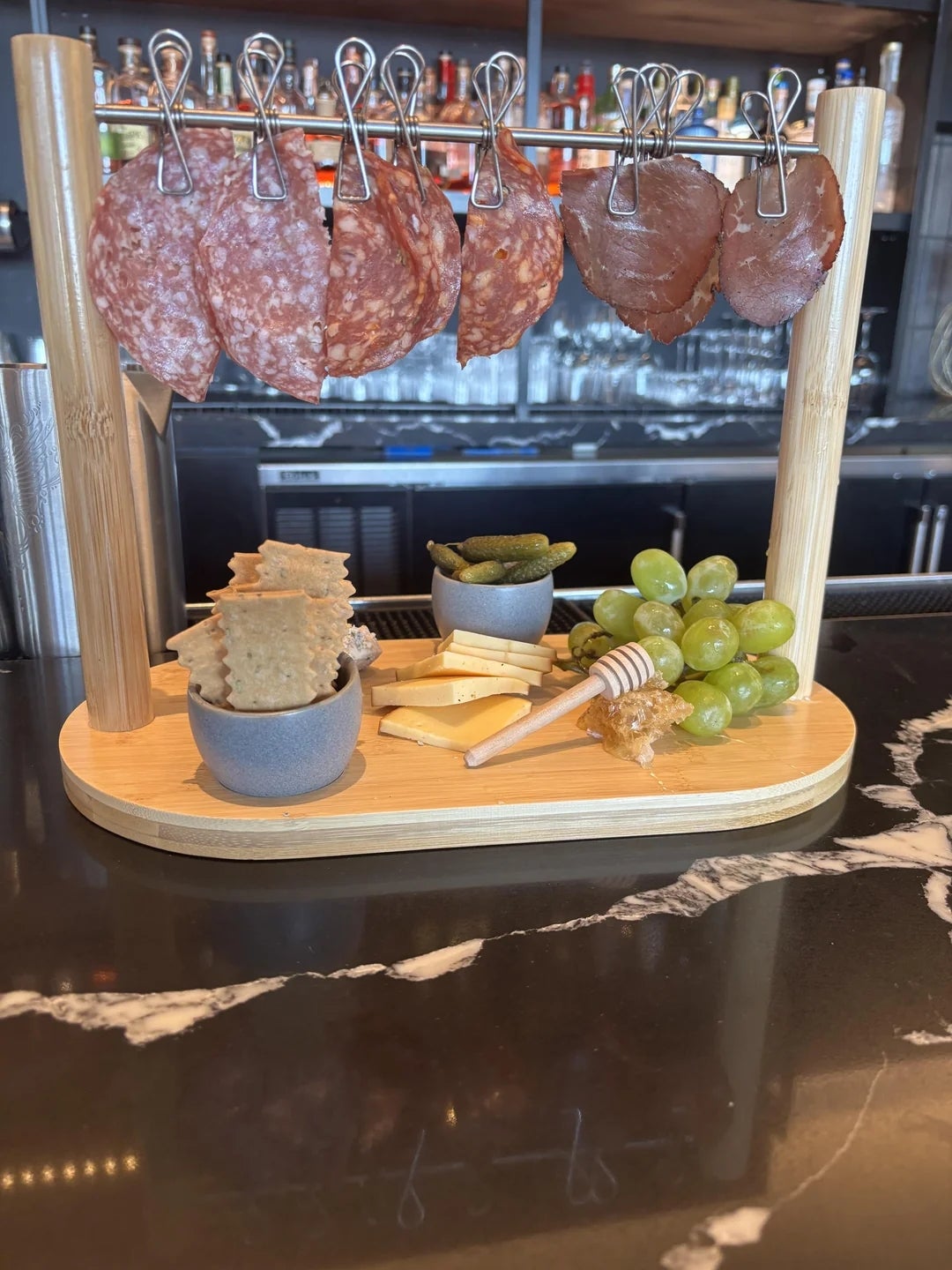 Charcuterie board with hanging salami, crackers, cheese, grapes, cornichons, and honey on a wooden board, set on a marble countertop