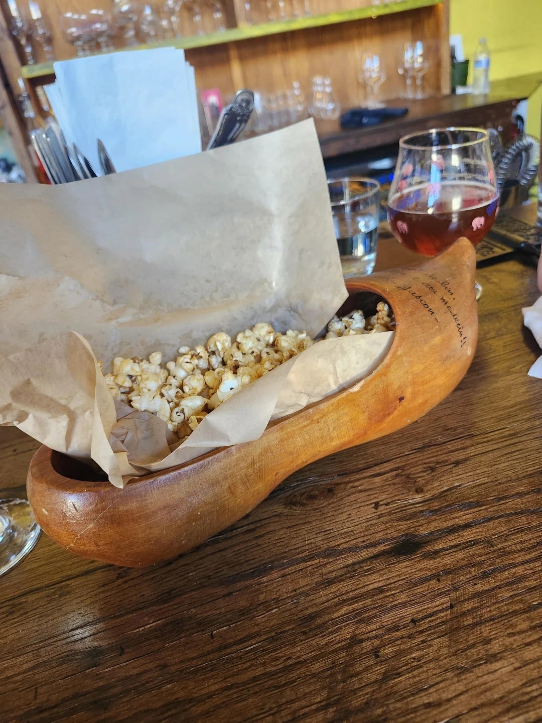 Popcorn in a wooden shoe-shaped bowl on a wooden table, next to a glass of wine