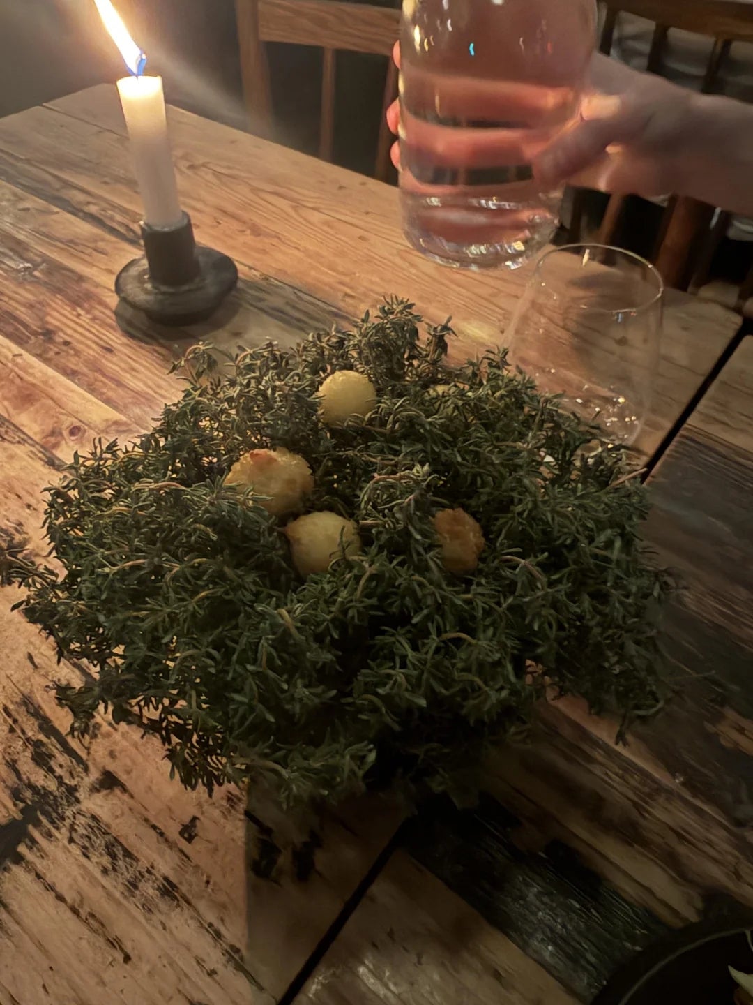 Dining table with a candle, a hand pouring water or wine, and a dish of crispy balls nestled in herbs