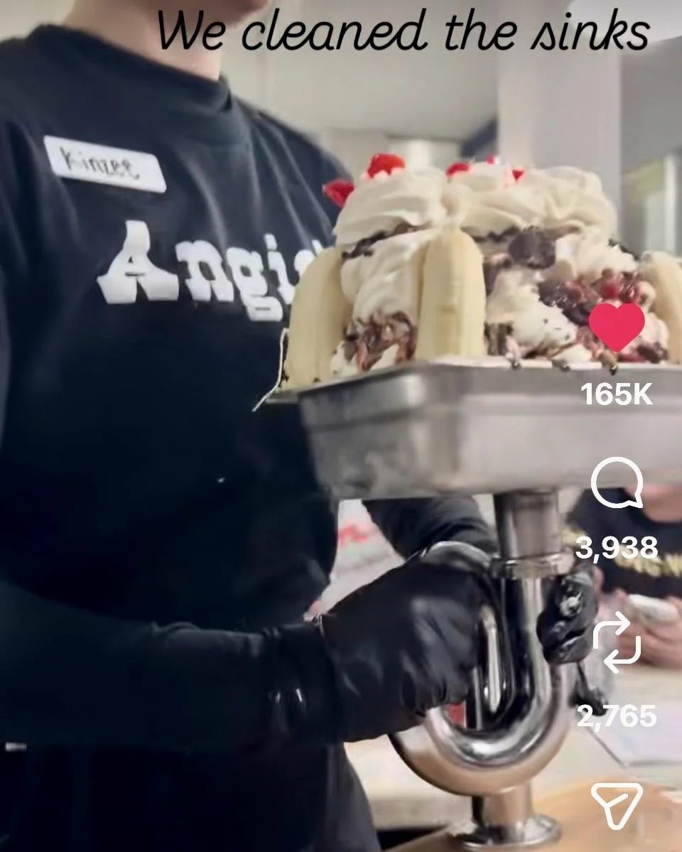 Person holding a massive banana split dessert in a metal dish shaped like a sink's plumbing. Text on image reads "We cleaned the sinks"