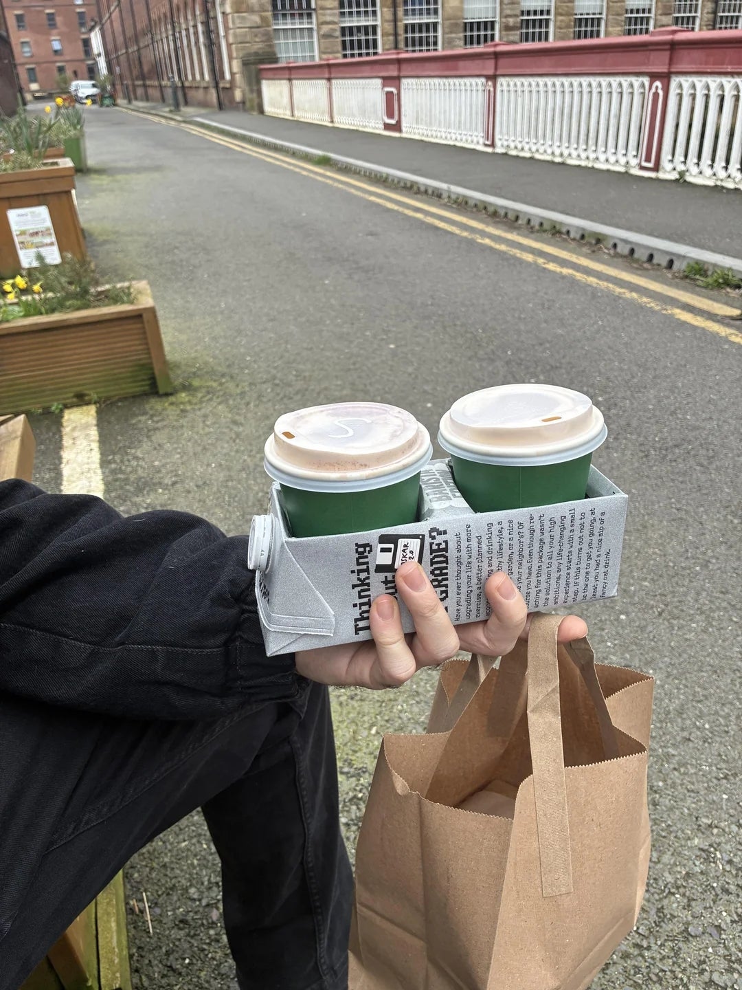 A person holds a brown paper bag and a coffee tray with two cups. They're sitting outdoors by a road