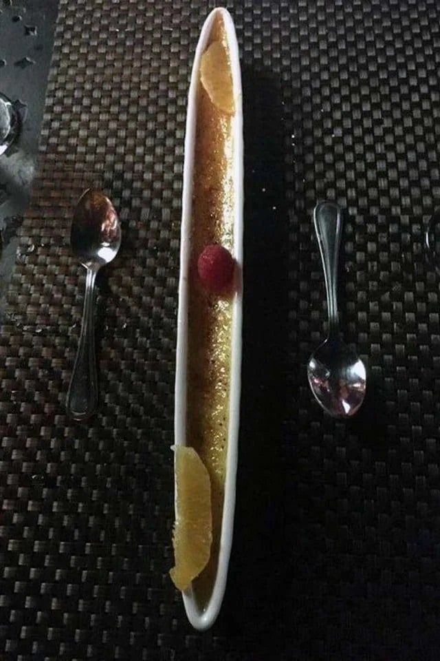 Long, narrow dessert dish with a golden surface, topped with a raspberry and citrus slices. Two spoons placed on a textured mat beside it