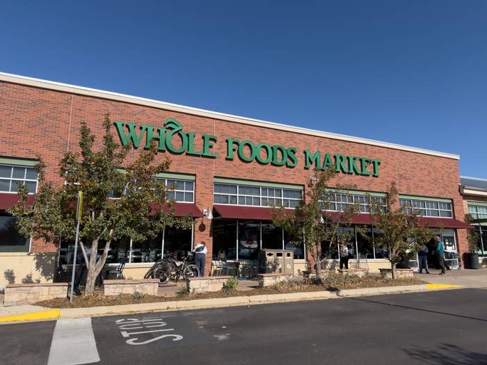 Whole Foods Market storefront with several shoppers outside, trees in the foreground, and a clear sky