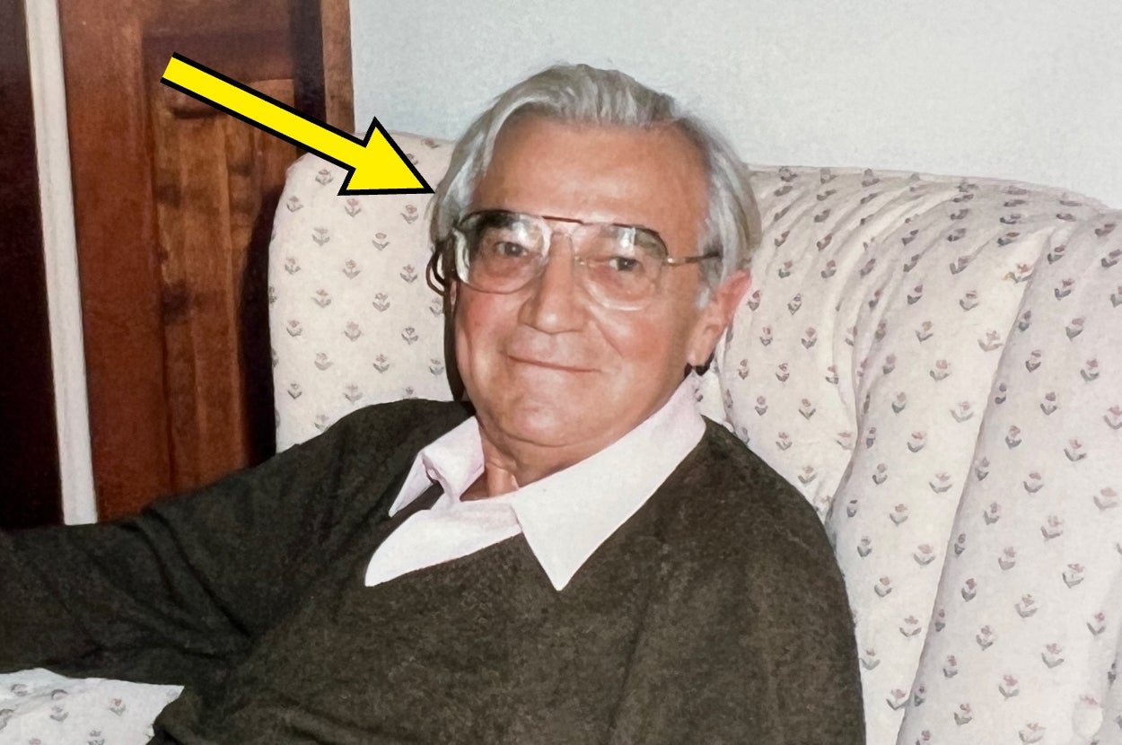 Person sitting on a patterned chair, wearing glasses and a sweater over a collared shirt, looking at the camera with a relaxed expression
