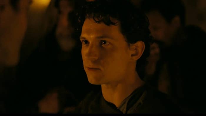 A young antheral   with curly hairsbreadth  appears focused successful  a dimly lit setting, surrounded by blurred figures successful  the background