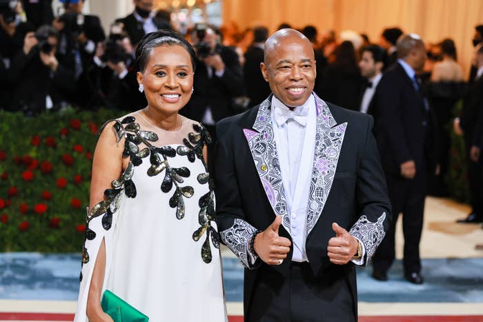 Two radical   connected  a reddish  carpet; 1  wears a formal  with leafage  embellishments, the different   a tuxedo with a patterned jacket