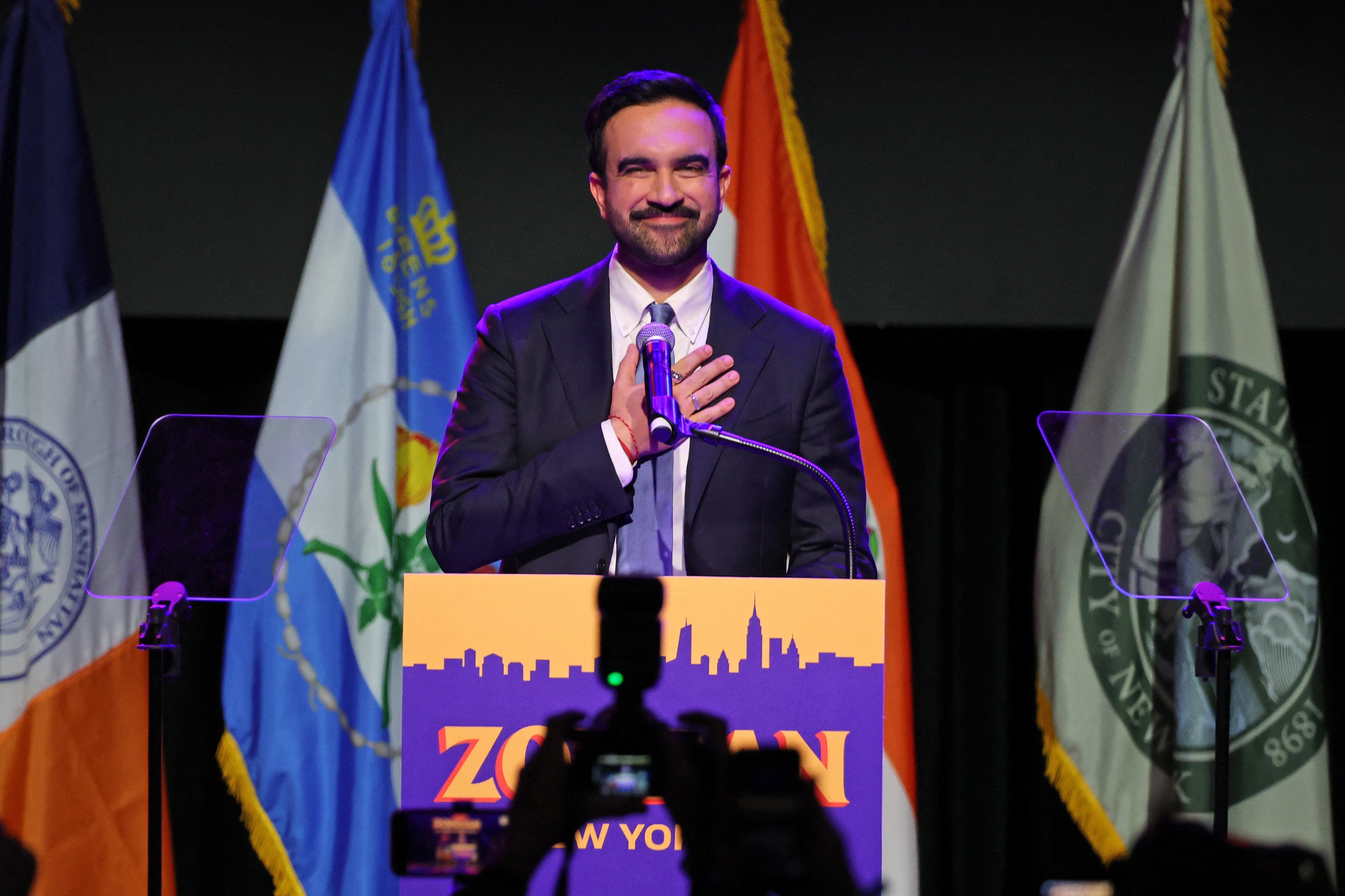 A antheral   successful  a suit   stands astatine  a podium with a sign, speaking to an audience. Various flags are displayed down  him