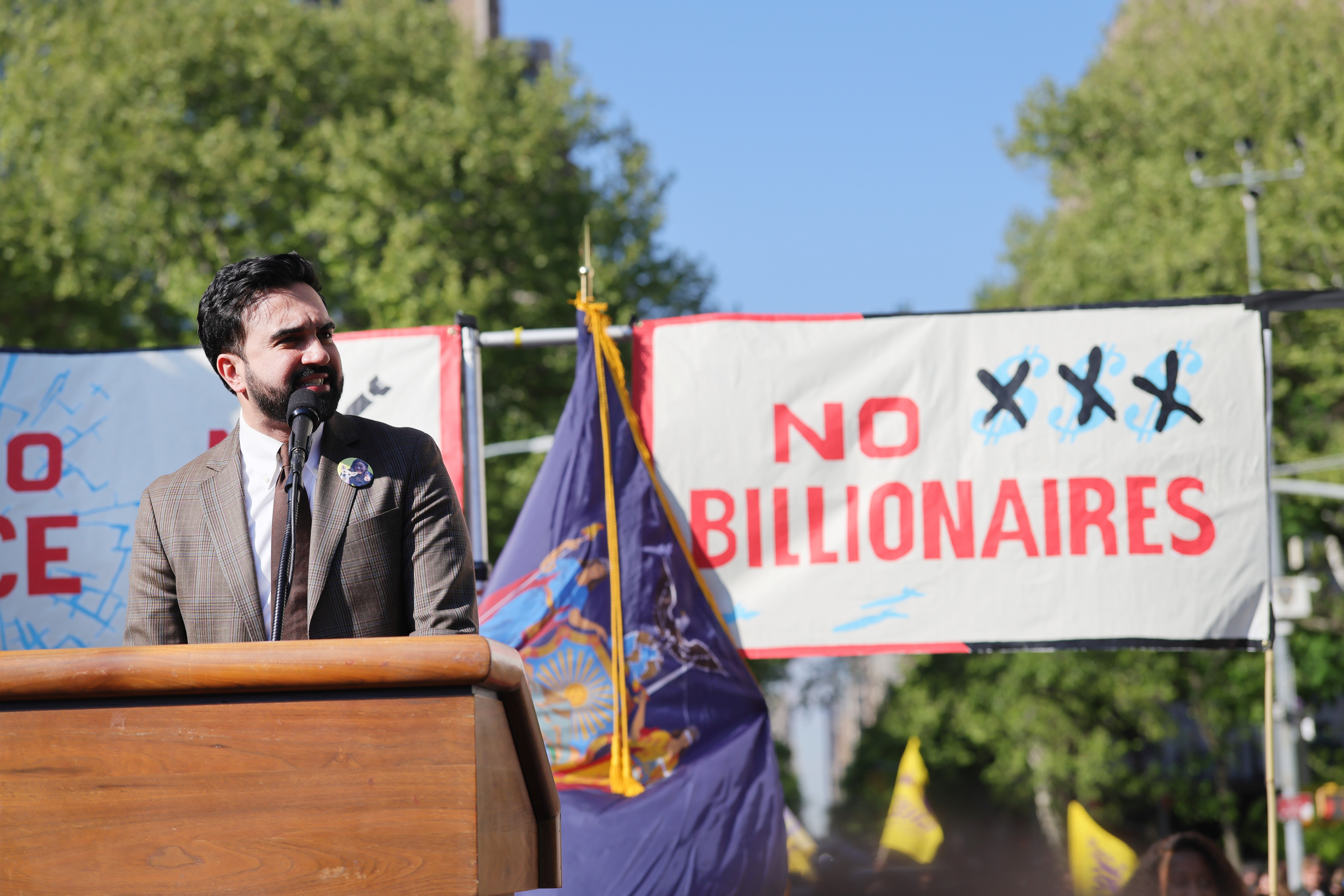 A idiosyncratic   astatine  a podium speaks astatine  a protestation  rally with signs saying "No Billionaires" successful  the background